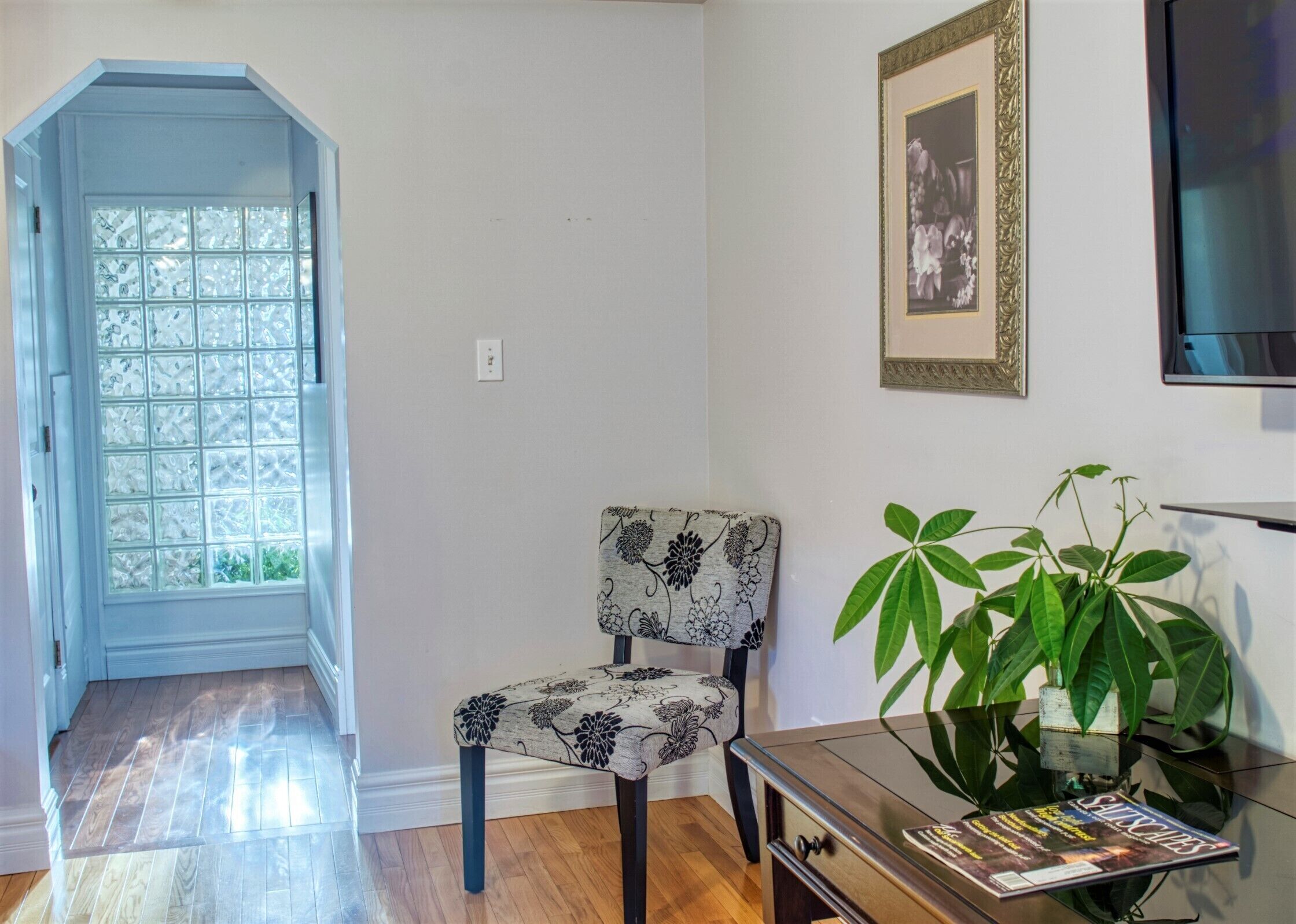 Una habitación con piso de madera, una silla con estampado floral, un cuadro en la pared y una planta sobre una mesa.
