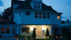 A two-story white house with a dark roof, a front porch with a lamp, and a small tree in the front yard. - Shamrock Suites (Florenceville-Bristol)