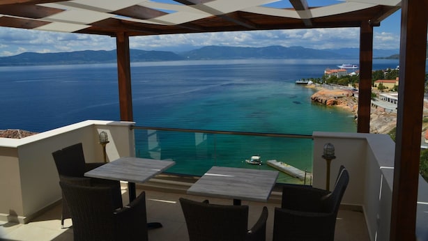 A covered outdoor seating area with tables and chairs, overlooking a clear blue sea and distant mountains. - Lydia Apartments (Istiaia-Aidipsos)