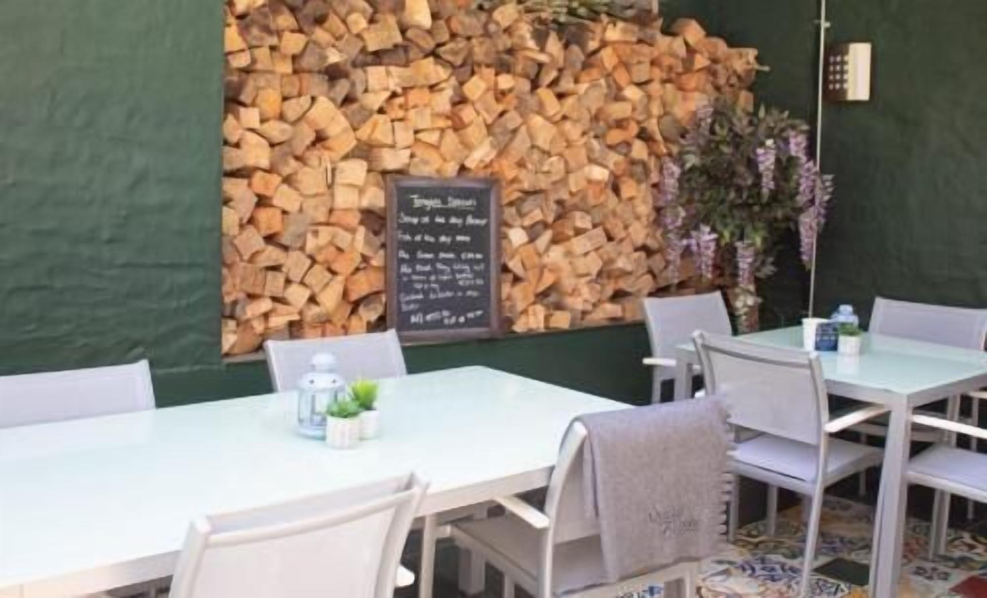 A restaurant with white tables and chairs, a stack of firewood against the wall, and a chalkboard menu.