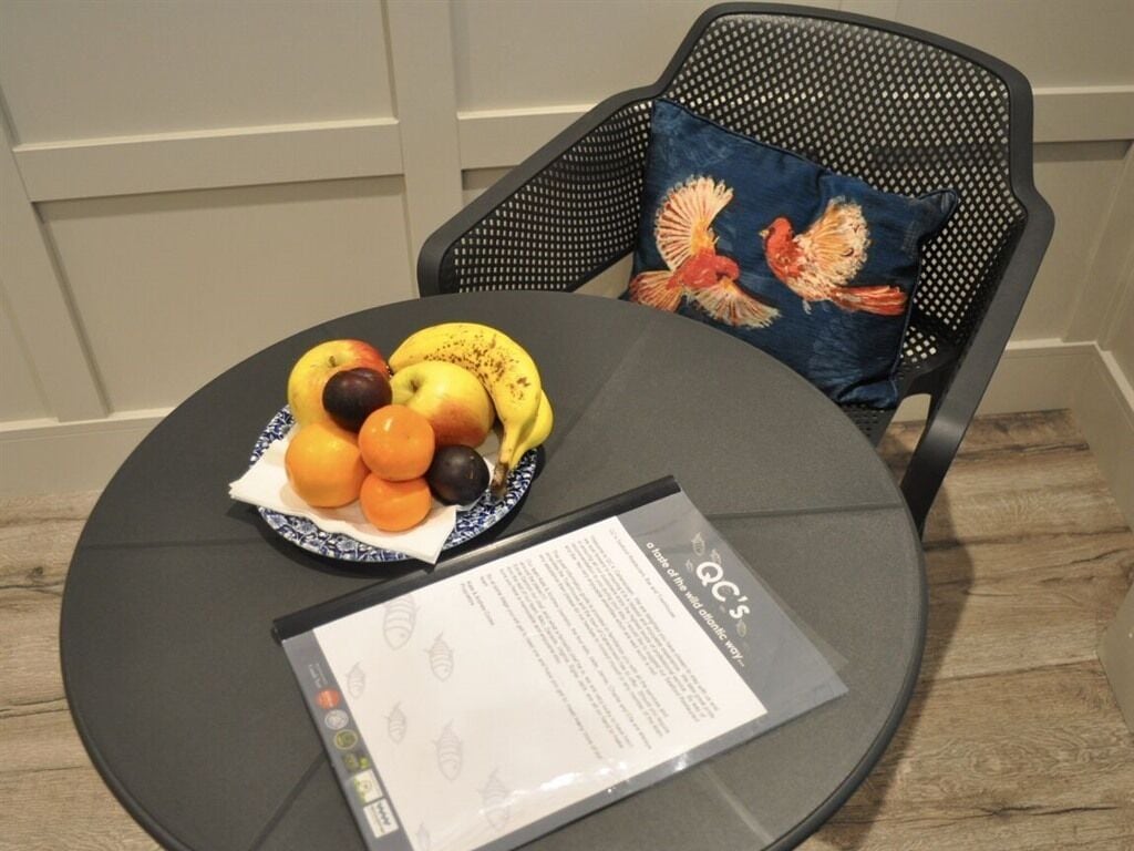 Une table ronde sur laquelle se trouve un saladier rempli de pommes, d'oranges et de bananes, un set de table avec du texte et une chaise agrémentée d'un coussin décoratif.