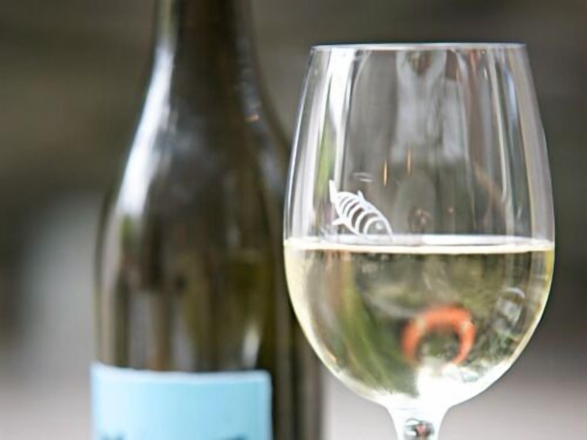 A wine glass with a clear liquid, possibly wine, and a bottle in the background.