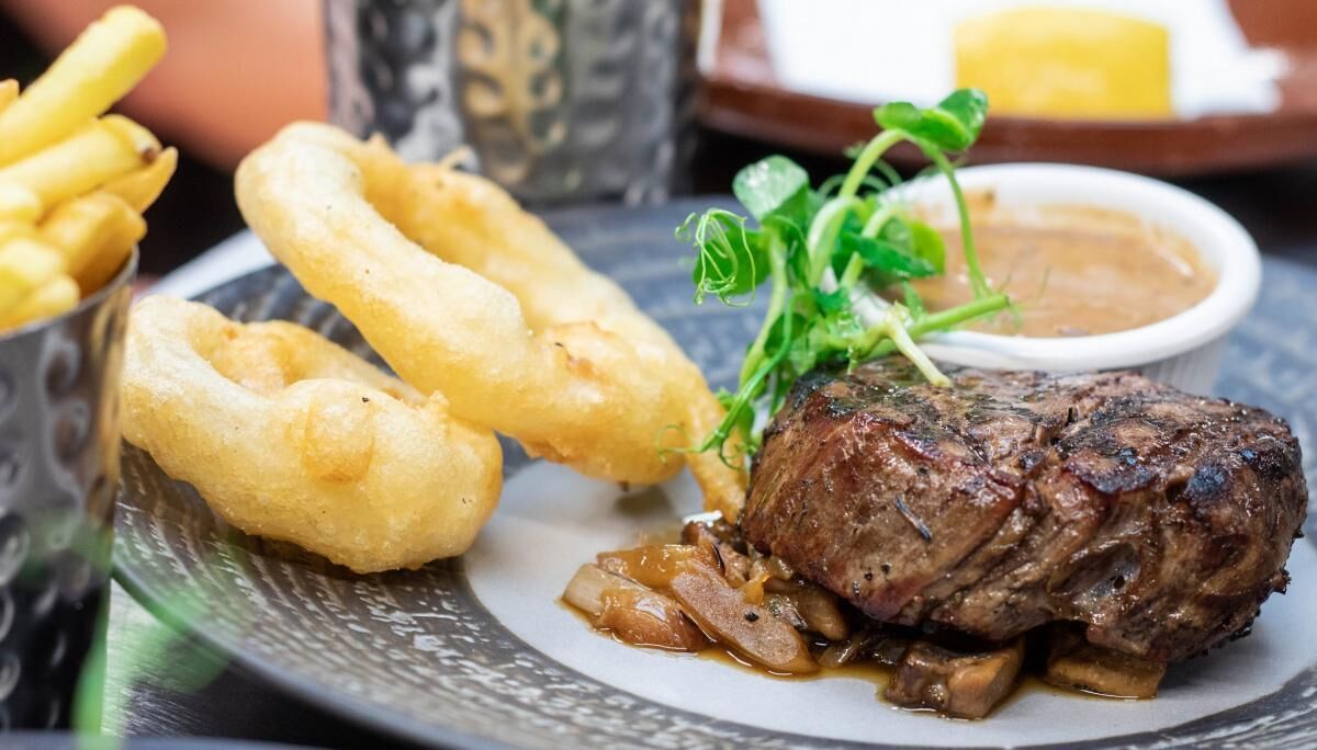 A plate with a steak, onion rings, and a small bowl of sauce.