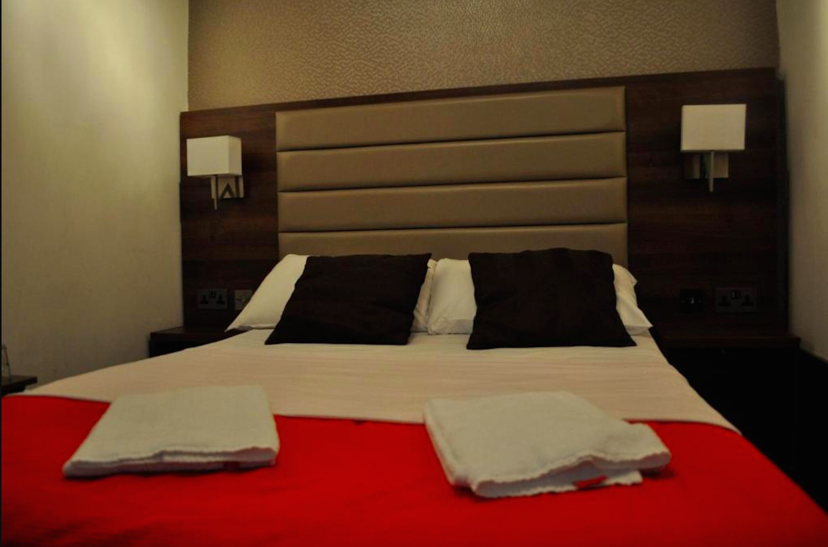 Una cama bien hecha con colcha roja y almohadas blancas, dos lámparas de noche y un cabecero de madera.