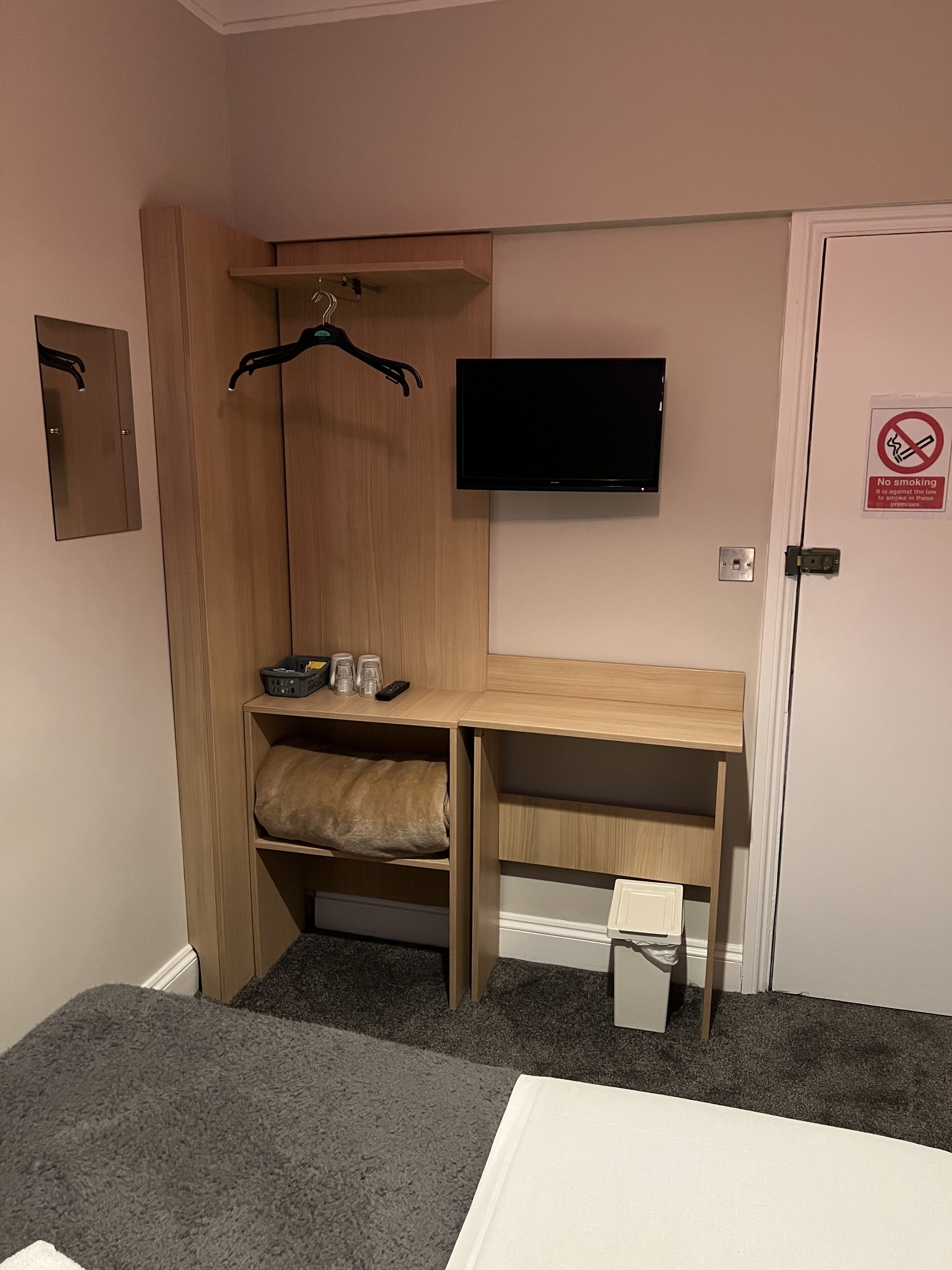 Una habitació petita amb un escriptori de fusta, un televisor fixat a la paret, un cartell de prohibició de fumar i una porta amb pany.