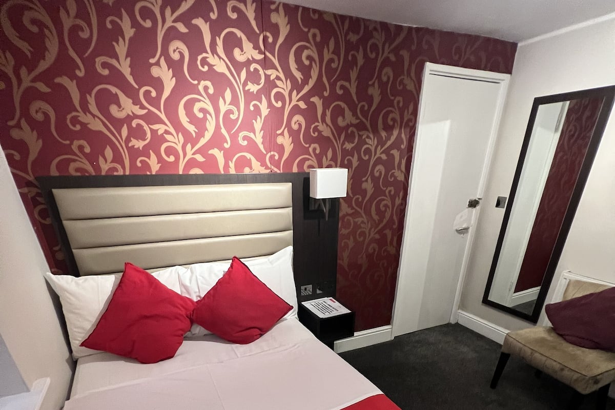 A hotel room with a large bed, red and white bedding, a nightstand with a lamp, a mirror, and a red chair.