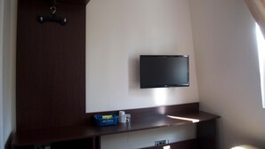A room with a flat-screen TV mounted on the wall, a wooden cabinet, and a small shelf with a basket and a glass. - Goodwood Hotel (London)