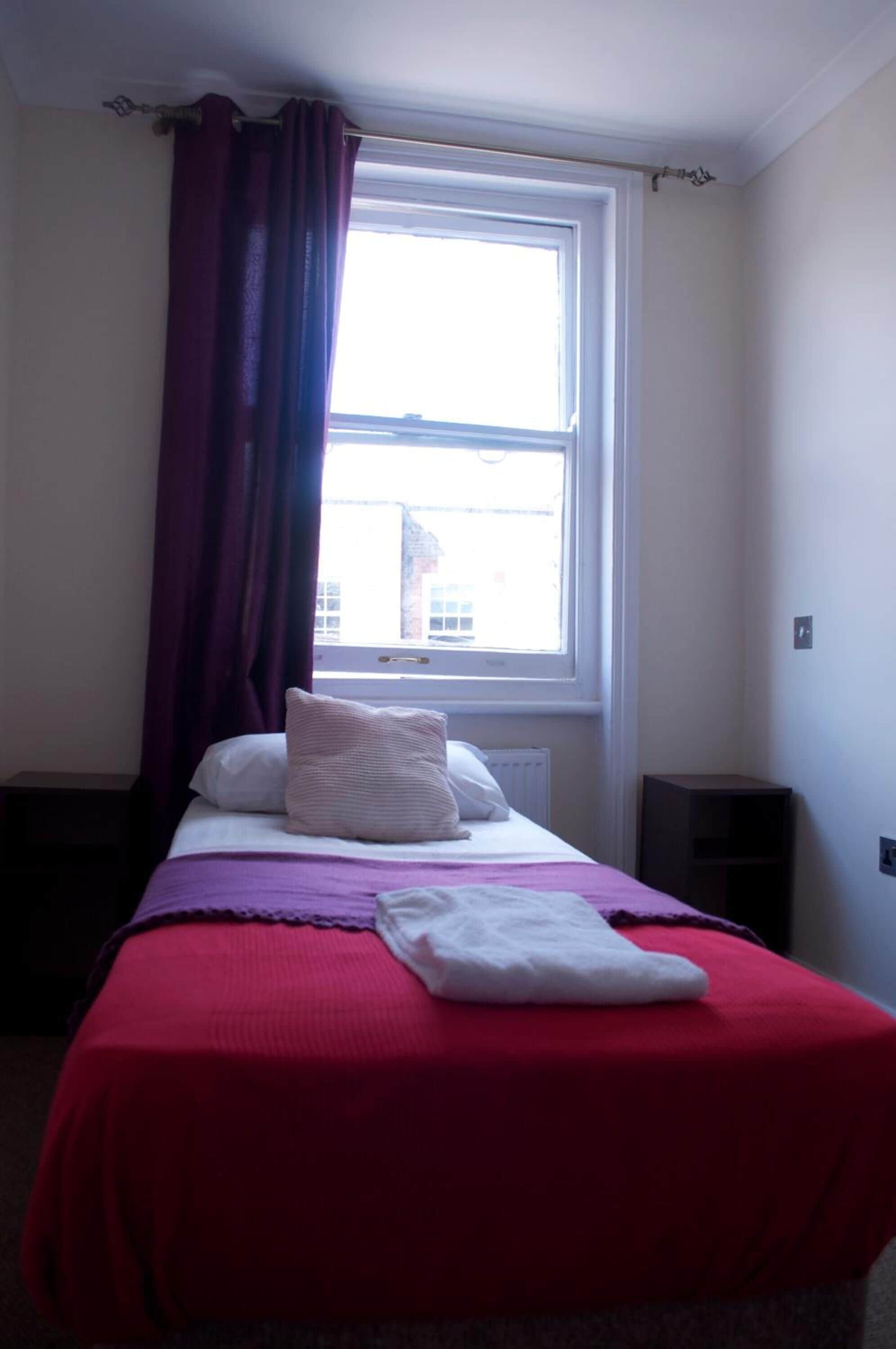 A neatly made bed with a red bedspread and white pillows, a window with a view of buildings, and a dark-colored nightstand beside the bed.