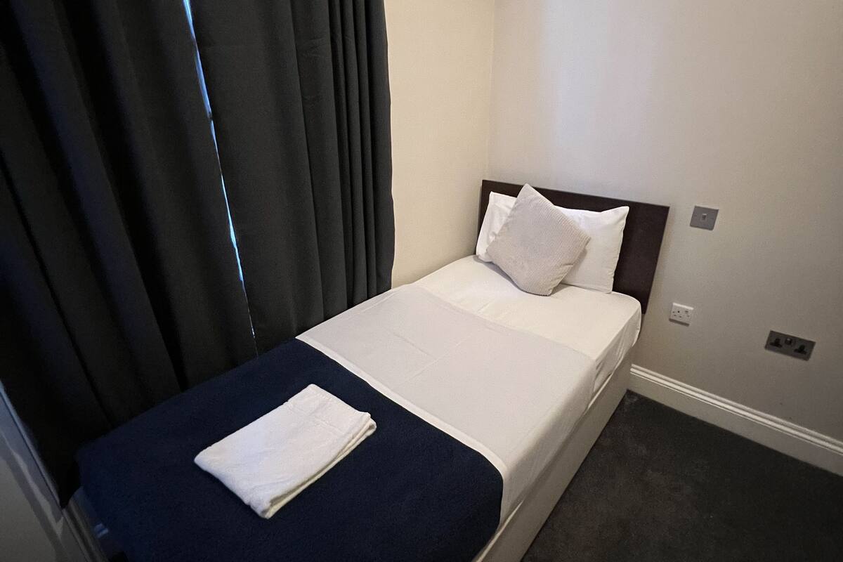 A single bed with a dark blue bedspread and white pillows, a white blanket, and a folded white towel on the bed.