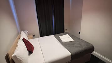 A neatly made bed with white and grey bedding, a wooden headboard, and a red pillow. There is a small wooden table beside the bed. The room has a dark carpet and a window with black curtains.