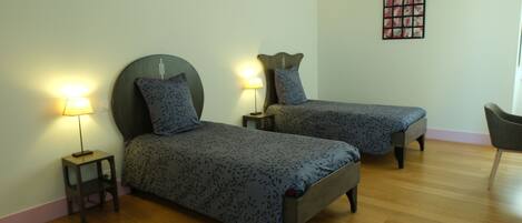 A room with two single beds, each with a nightstand and a lamp. The room has wooden flooring and a wall-mounted artwork.