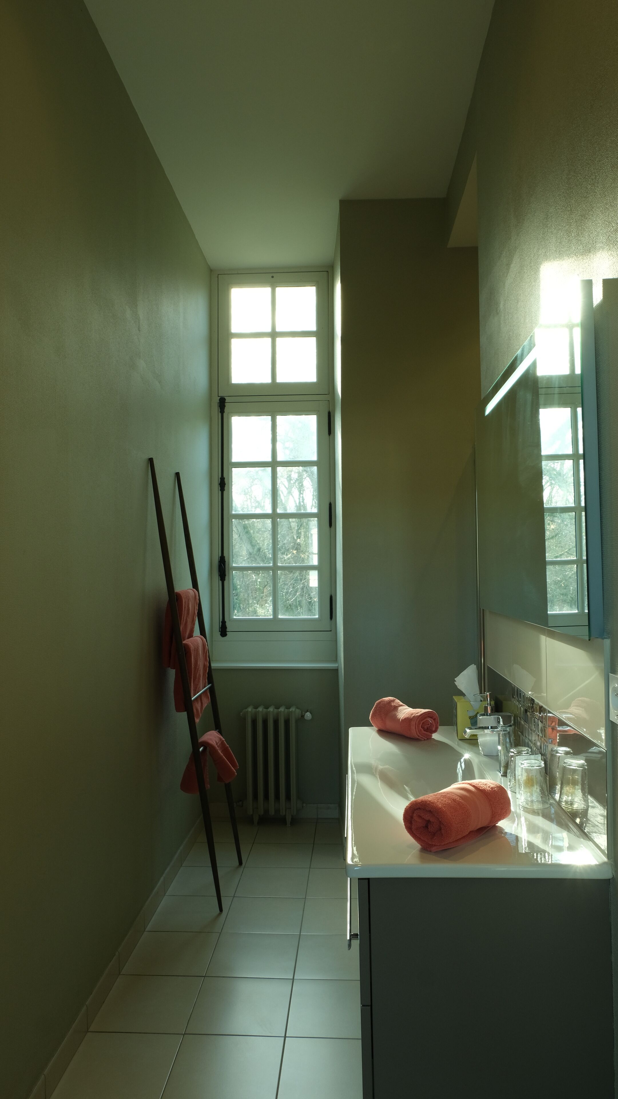 A bathroom with a window, a radiator, a towel rack, and a sink with glass jars.