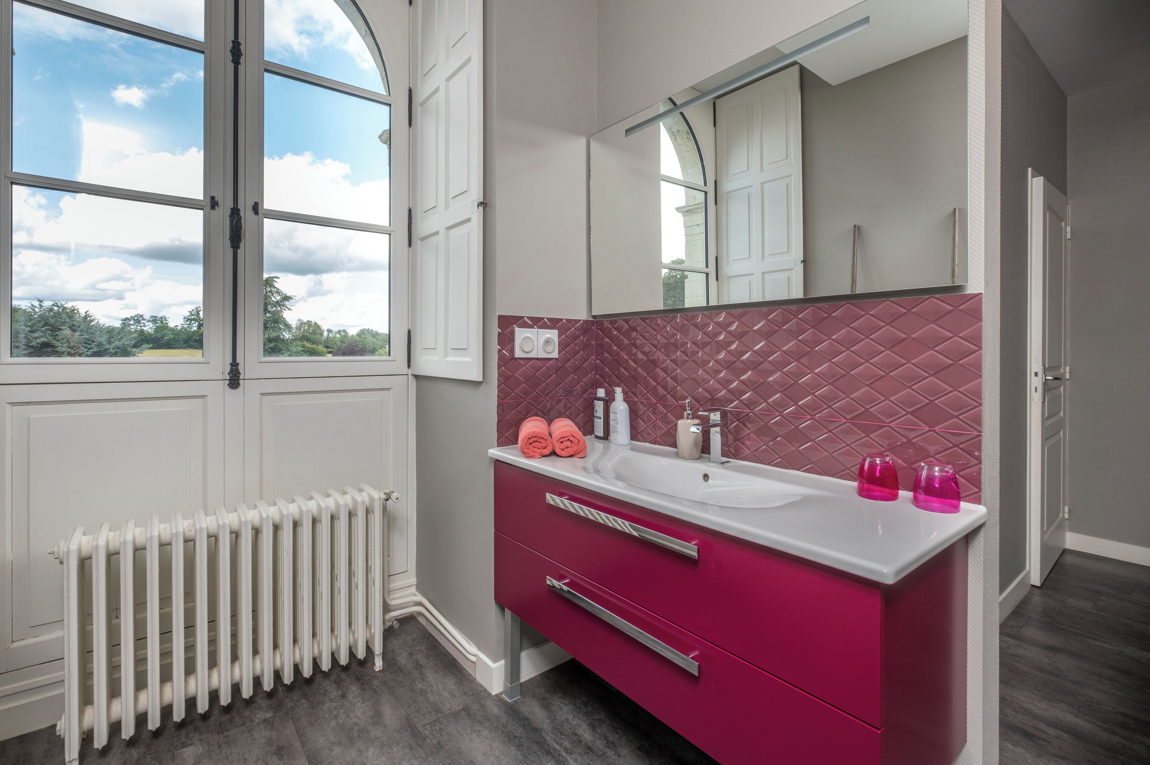 Ein Badezimmer mit großem Fenster, einem weißen Heizkörper, einem rosafarbenen Waschtisch und einer Rückwand mit Diamantmuster sowie einem Spiegel über dem Waschbecken.