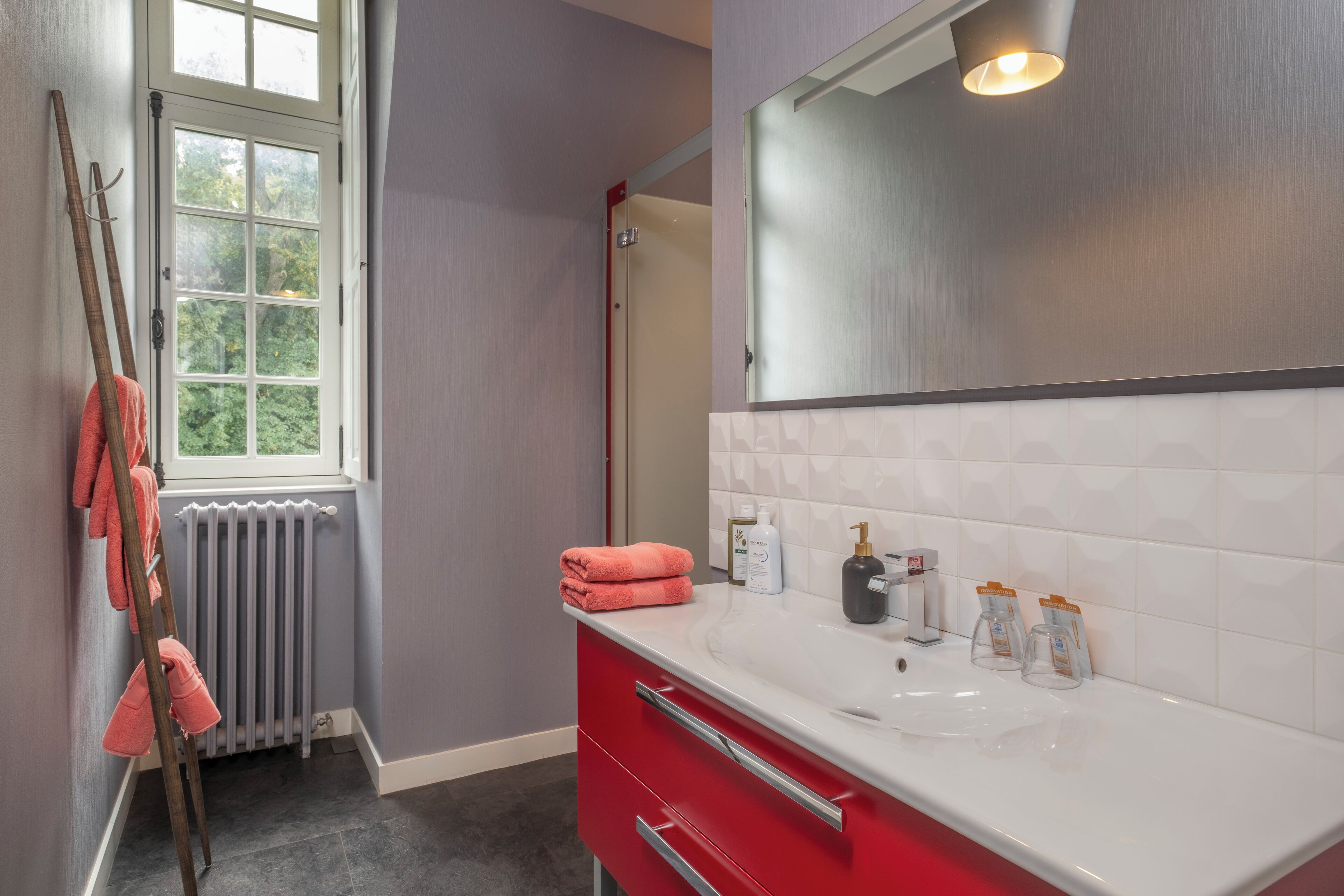 Une salle de bain avec un lavabo blanc, un meuble rouge et une fenêtre donnant sur des arbres.