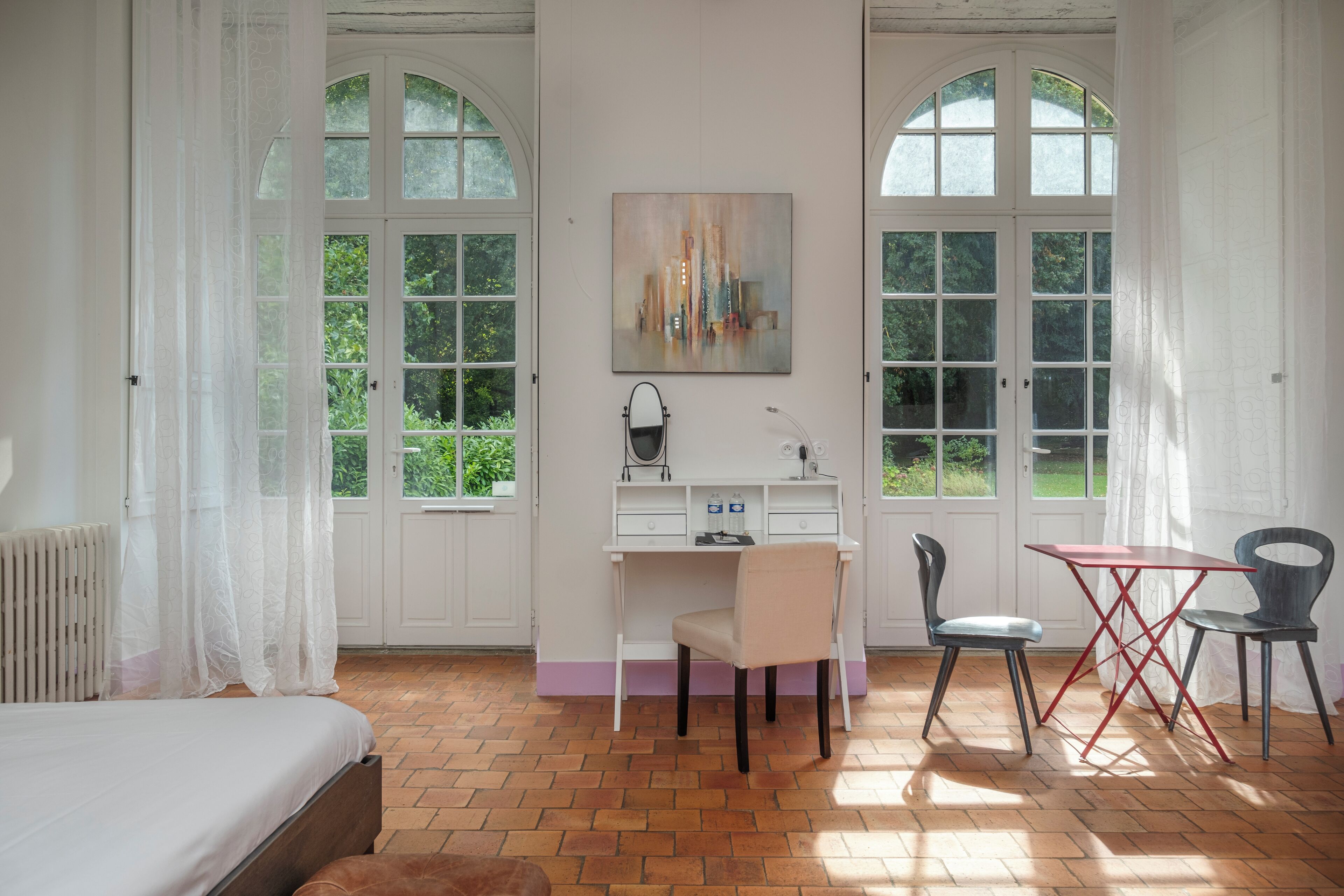 A bedroom with a bed, a desk, two chairs, and a window with a view of greenery.