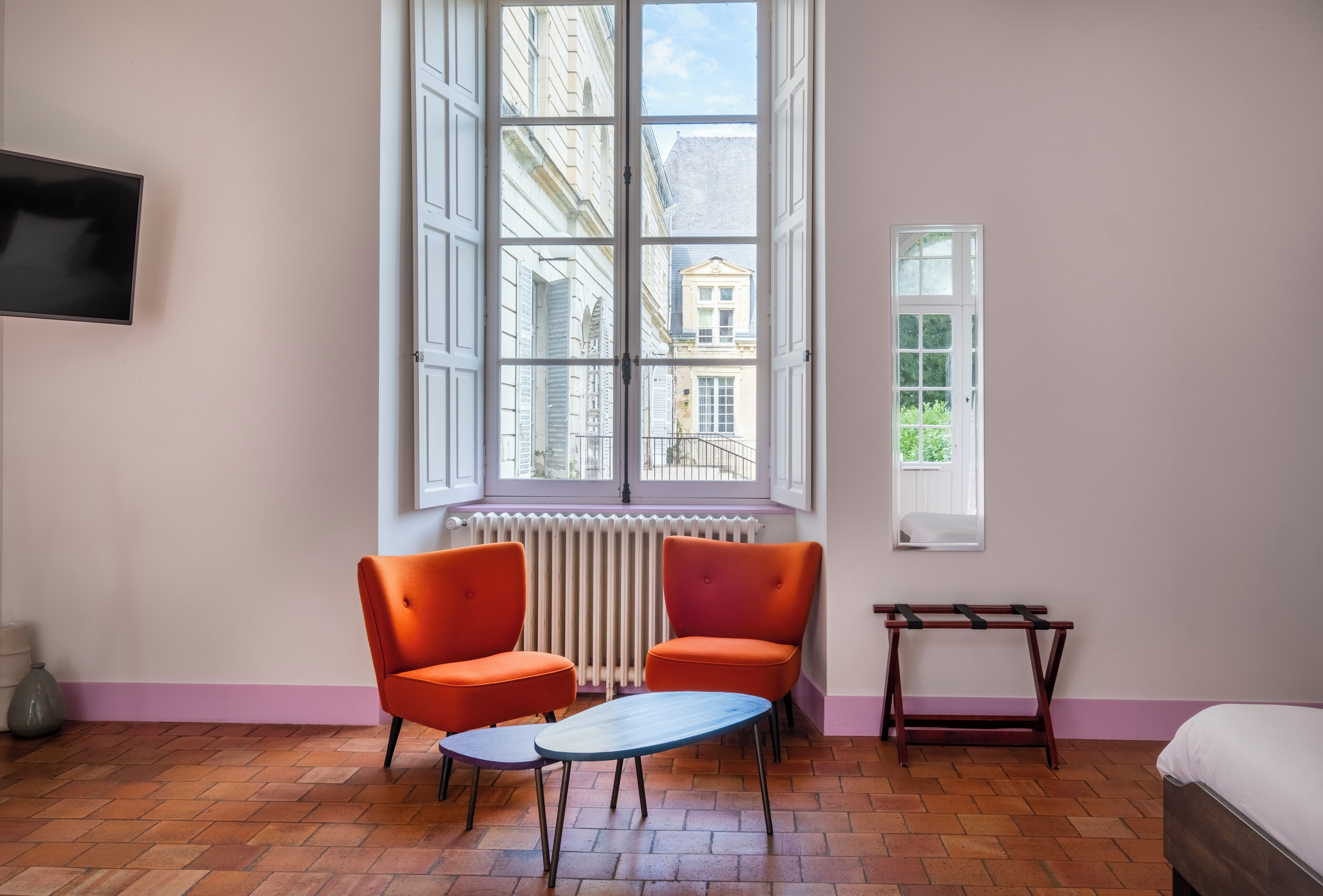 A room with two orange chairs, a small table, a bed, a radiator, and a window with a view of buildings.