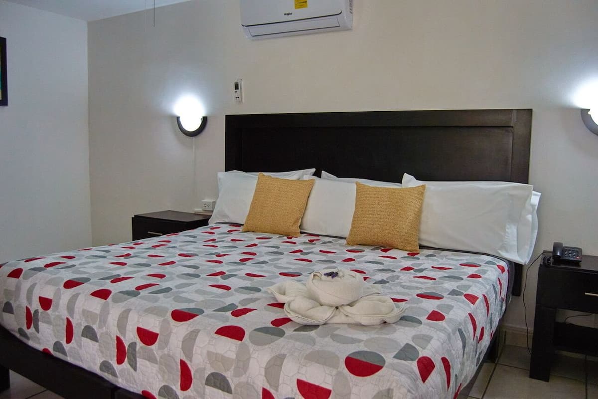 A hotel room with a bed, two bedside tables, a wall-mounted air conditioner, and a wall-mounted light fixture.