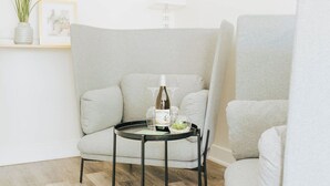A corner of a room with a grey armchair, a small round table with a bottle and glasses, and a framed picture on a shelf. - Naramata Courtyard Suites (Naramata)