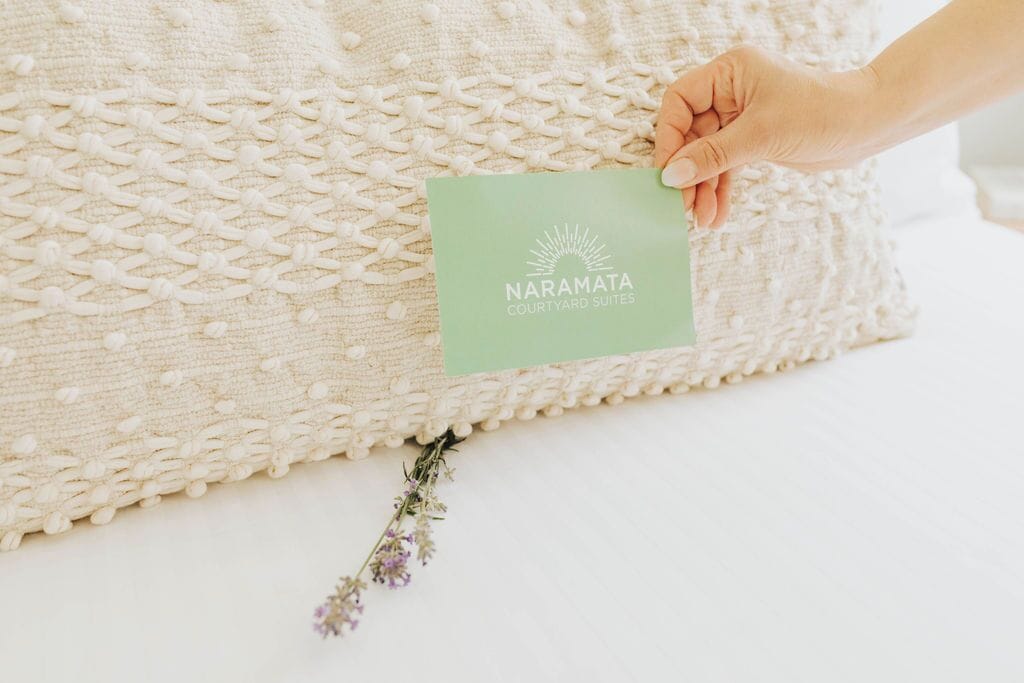 Una mano sostiene una tarjeta verde con el texto "NARAMATA COURTYARD SUITES", colocada sobre un almohadón beige con textura.