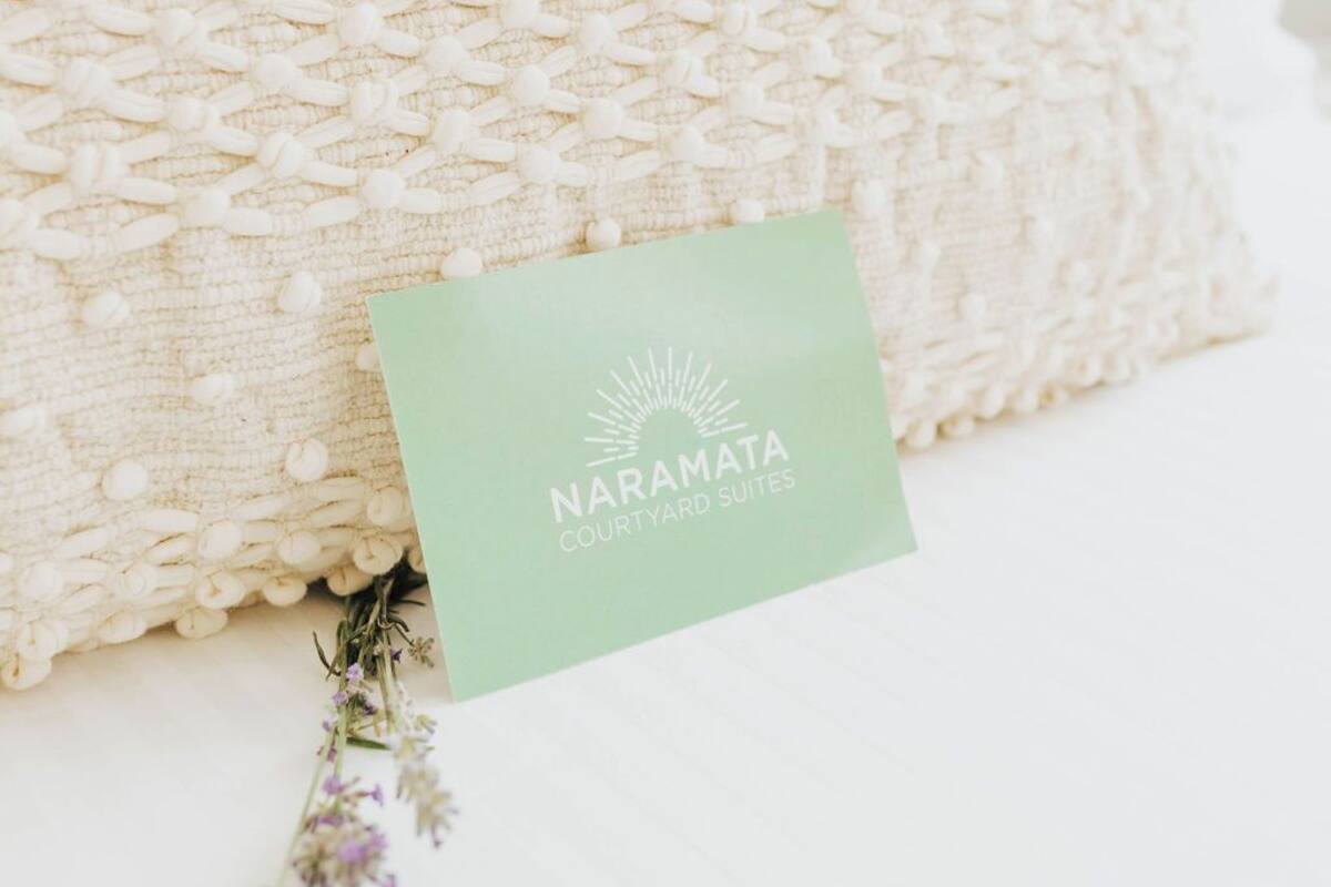 A white textured pillow with a green card labeled "NARAMATA COURTYARD SUITES" and a small bunch of purple flowers.