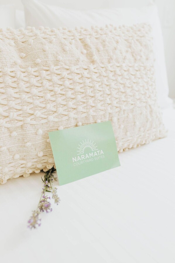 A white textured pillow with a green card labeled "NARAMATA COURTYARD SUITES" and a small bunch of purple flowers. - Naramata Courtyard Suites (Naramata)