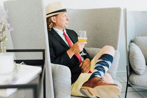 A man in a suit and hat sitting in a grey armchair, holding a glass of beer. - Naramata Courtyard Suites (Naramata)
