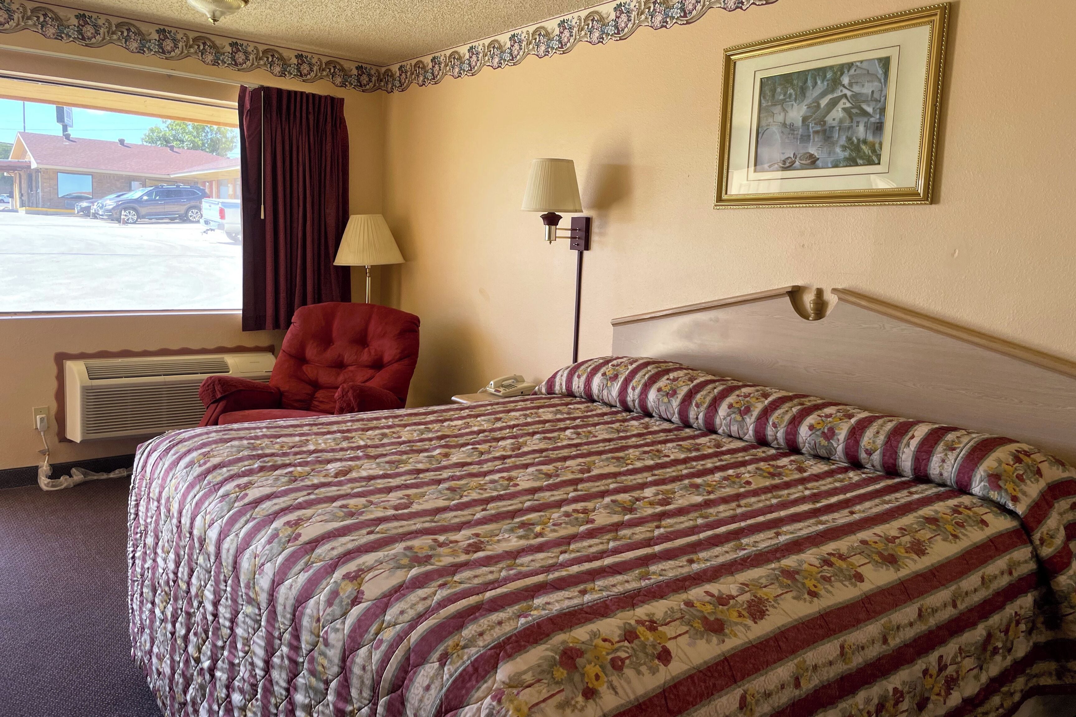 Une chambre d’hôtel avec un grand lit, un fauteuil rouge, une table de chevet avec une lampe, un tableau encadré au mur et une fenêtre donnant sur un immeuble et des voitures garées à l’extérieur.