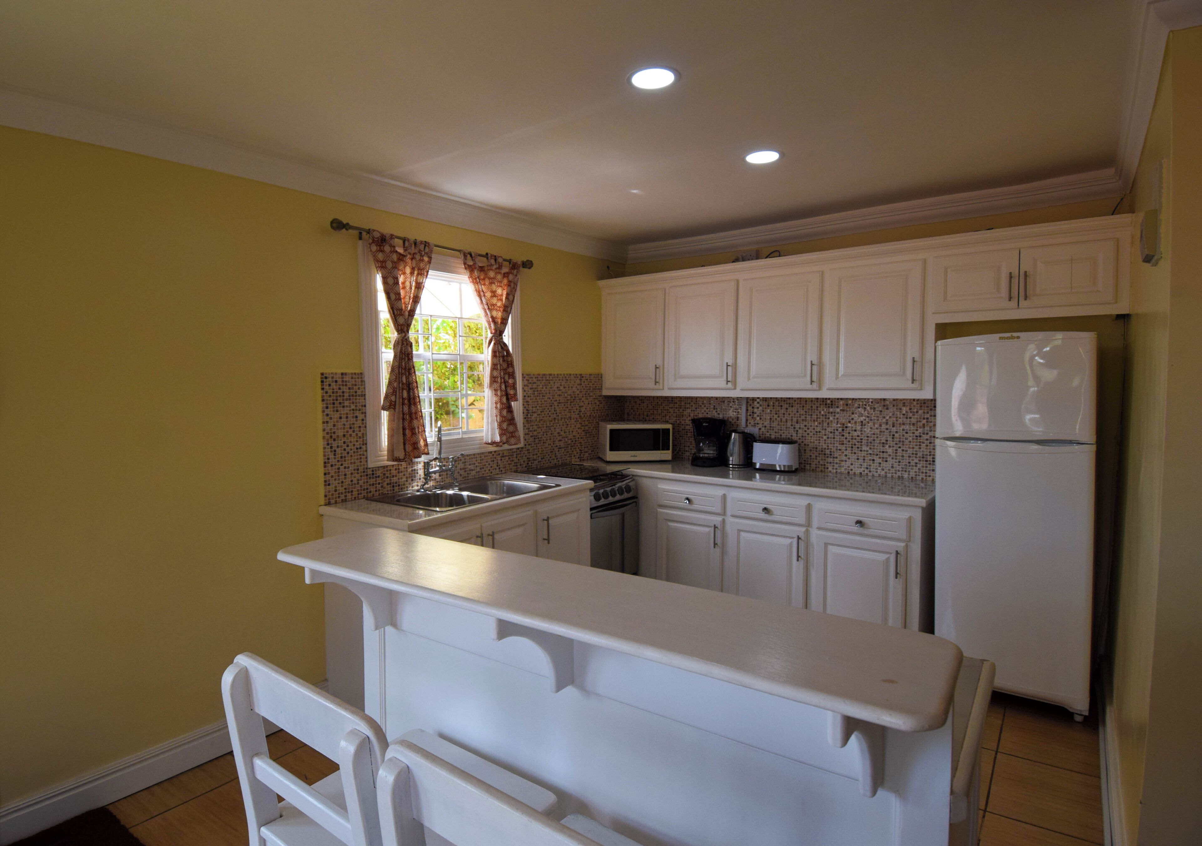 A compact kitchen with white cabinets, a microwave, a refrigerator, and a window with curtains.