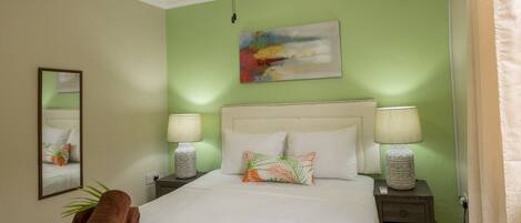 A neatly made bed with white linens, two bedside lamps, a ceiling fan, and a painting on the wall.