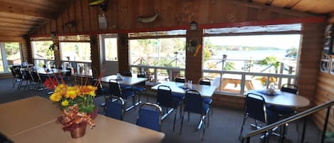 A wooden cabin-style restaurant with tables and chairs, large windows offering a view of the lake, and a flower arrangement on the table.