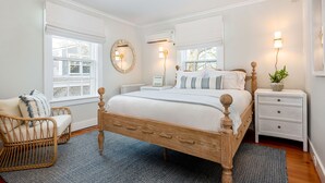 A bedroom with a wooden bed, a wicker chair, a bedside table with a lamp, and a window with white curtains.