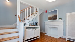 A staircase with wooden steps and white railings, a white console table with a plant, a white fireplace, and a framed picture on the wall. - Periwinkle Inn (Nantucket)