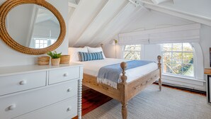 A bedroom with a wooden bed, white dresser, and a large mirror. There is a window with a view of trees and a white ceiling with exposed beams.