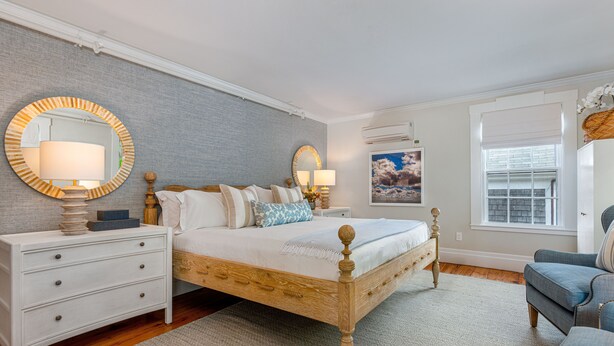 A bedroom with a wooden bed, white nightstands, a round mirror, and a window with blinds.