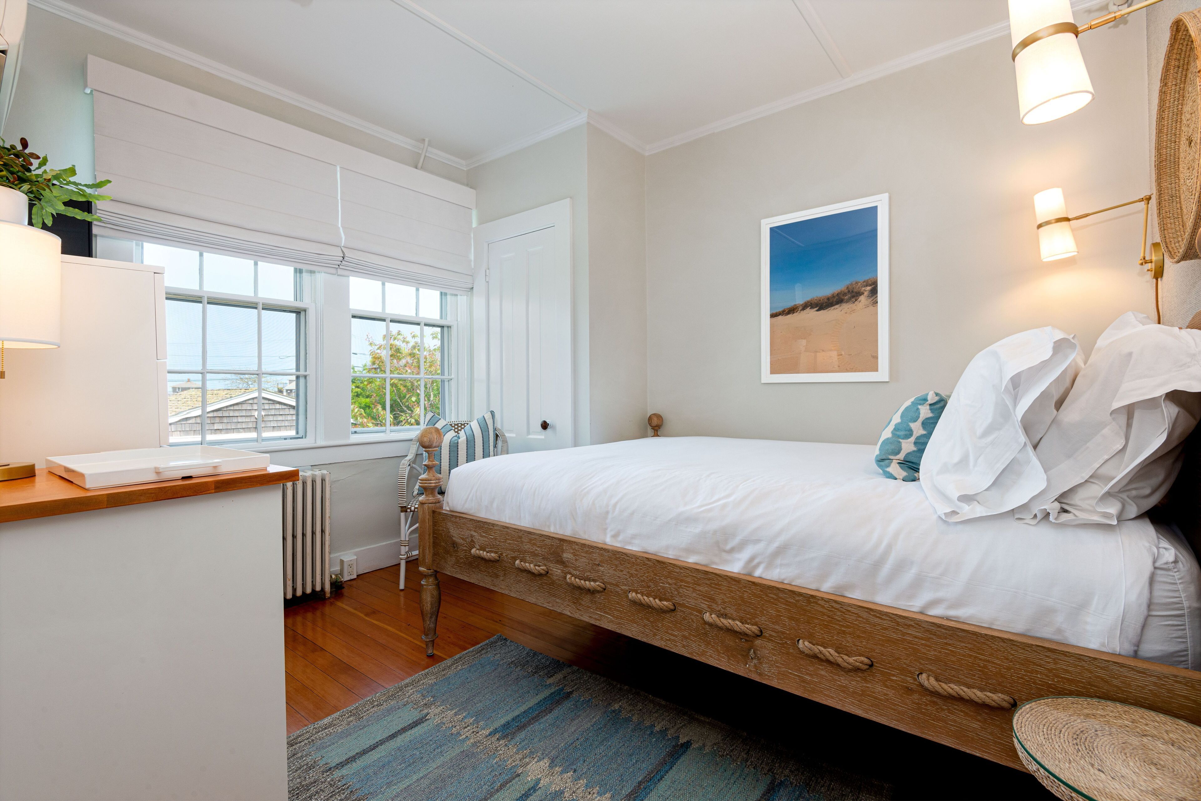 A bedroom with a wooden bed, white bedding, a view of the sea through a window, a wall-mounted lamp, and a framed picture.