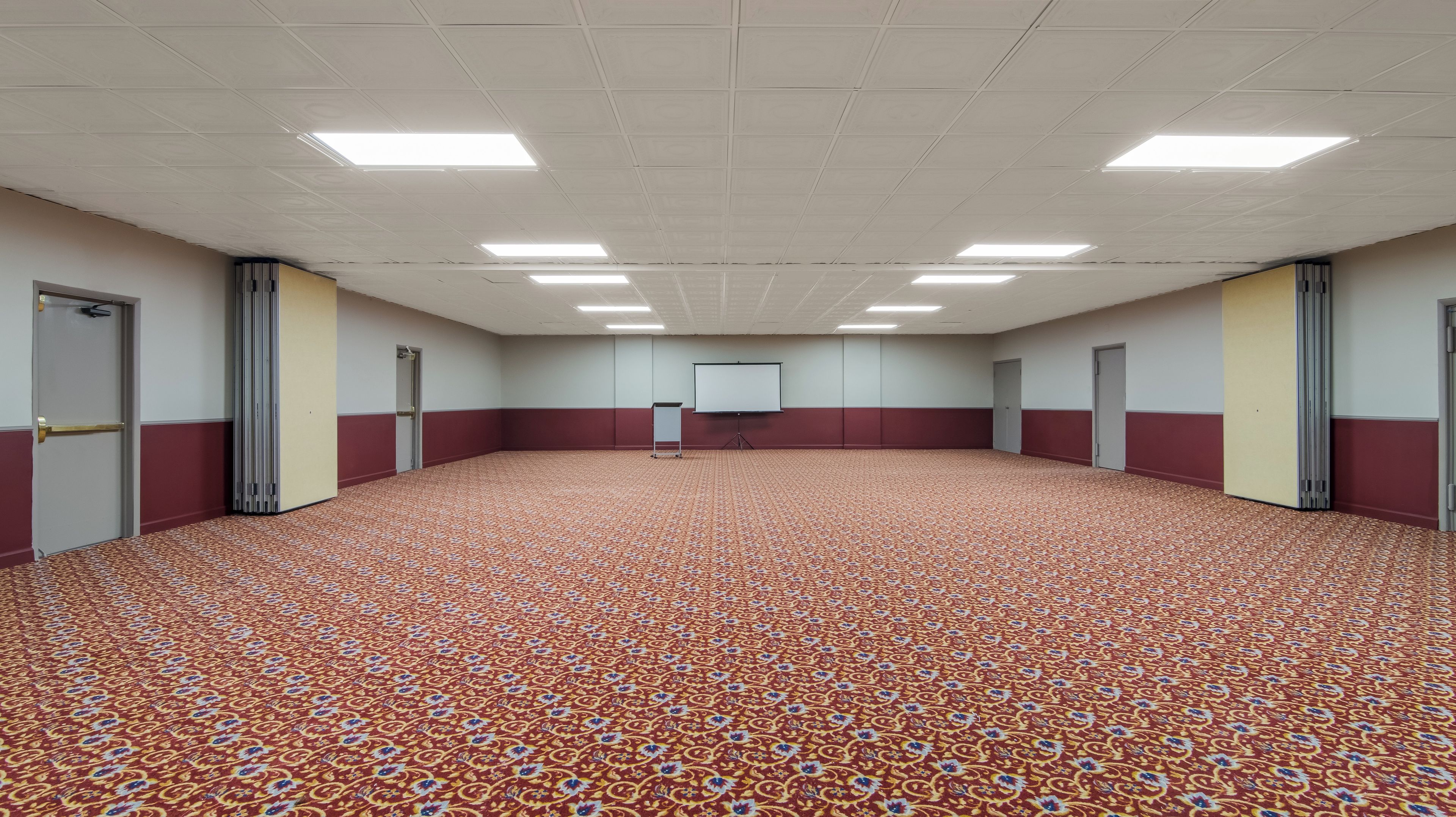 A large conference room with a patterned carpet, a white projection screen, and doors along the walls.