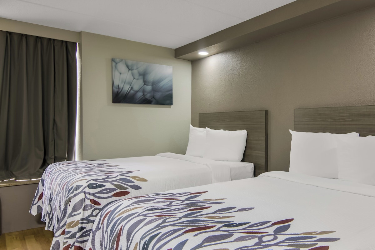 A hotel room with two beds, a large window with curtains, a painting on the wall, and a wooden headboard.