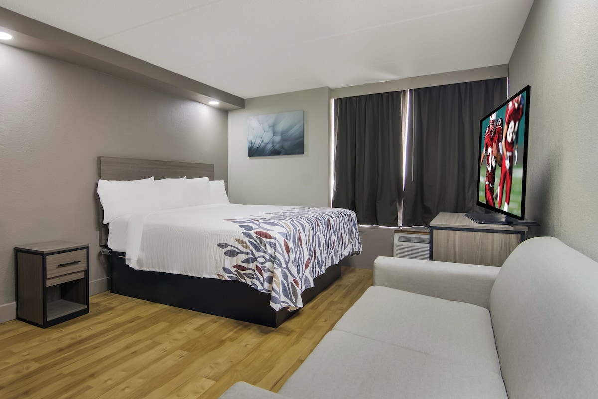 A hotel room with a bed, a bedside table, a TV, a couch, and a window with curtains.