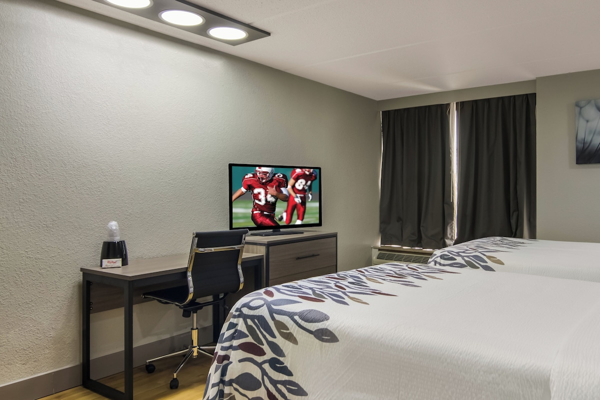 A hotel room with a bed, a desk with a chair, a television displaying a sports game, a window with curtains, and a painting on the wall.