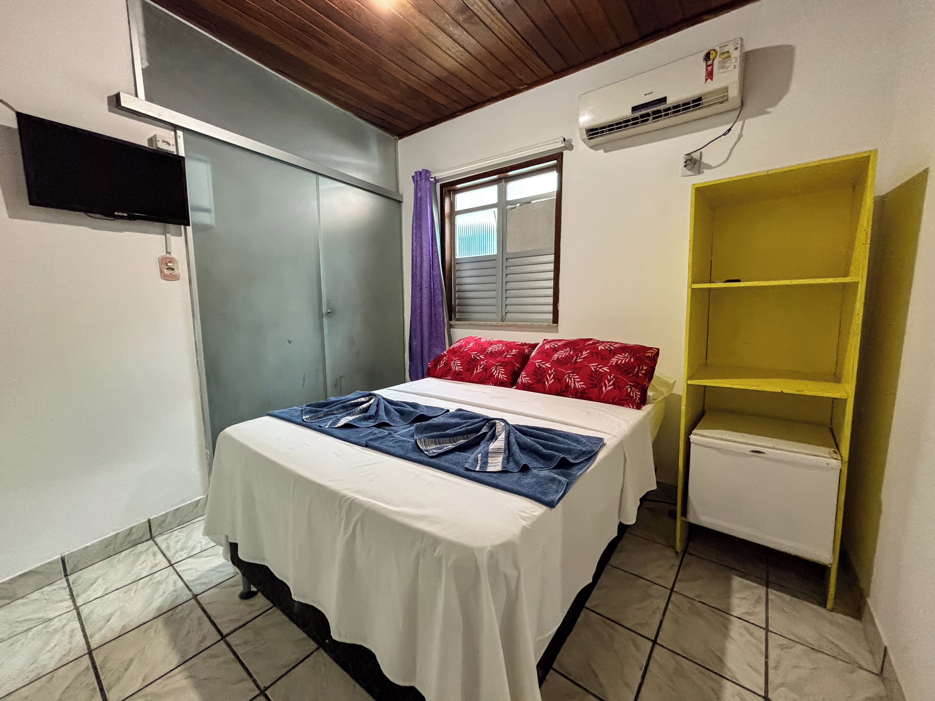 A small bedroom with a bed, a wardrobe, a TV, an air conditioner, and a yellow shelf.