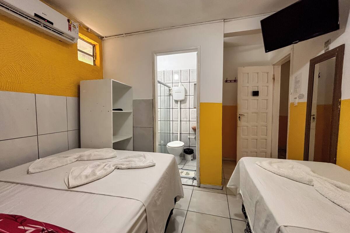 Habitación de hotel con dos camas, un televisor, un equipo de aire acondicionado, un baño con inodoro y lavamanos, y una puerta que da a otra habitación.