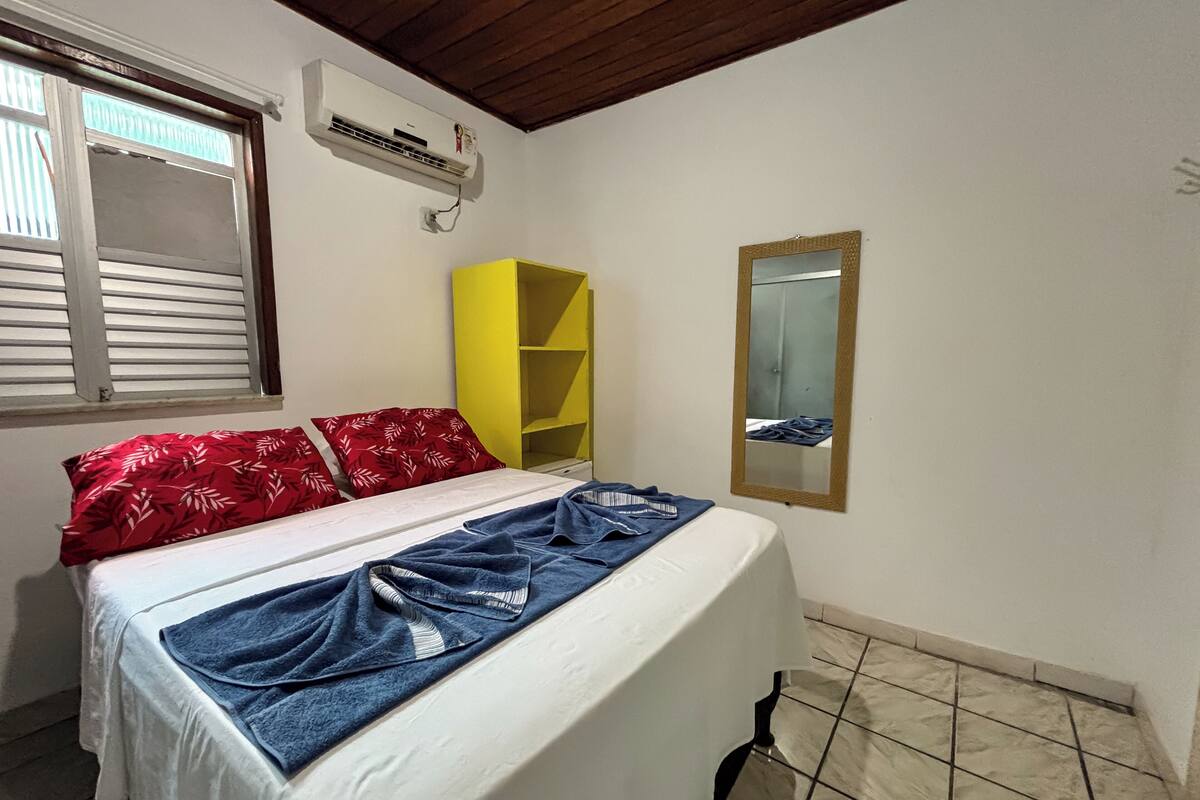 Un dormitorio con cama, ventana con persianas, un estante amarillo y un espejo en la pared.