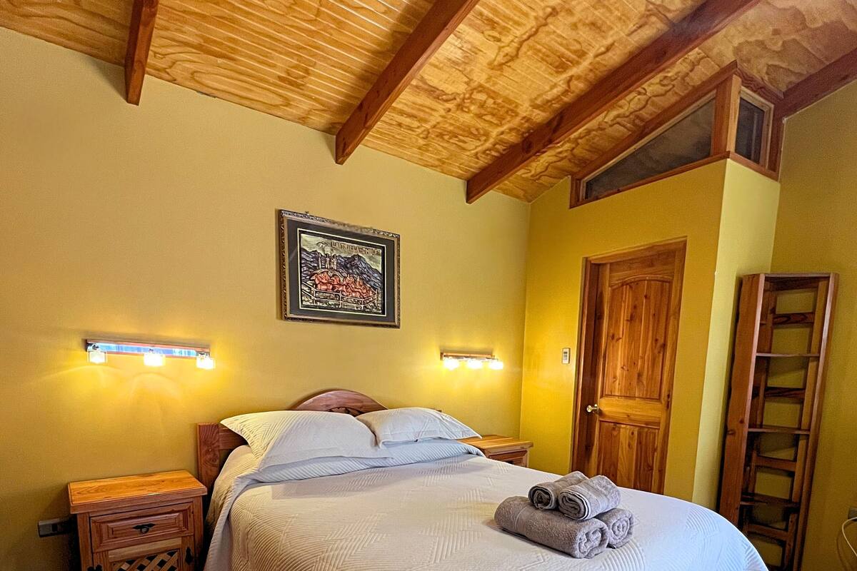 Un lit bien fait, avec une couette blanche et des oreillers, une table de chevet en bois avec un tiroir, une tête de lit en bois, un tableau accroché au mur et une porte en bois.
