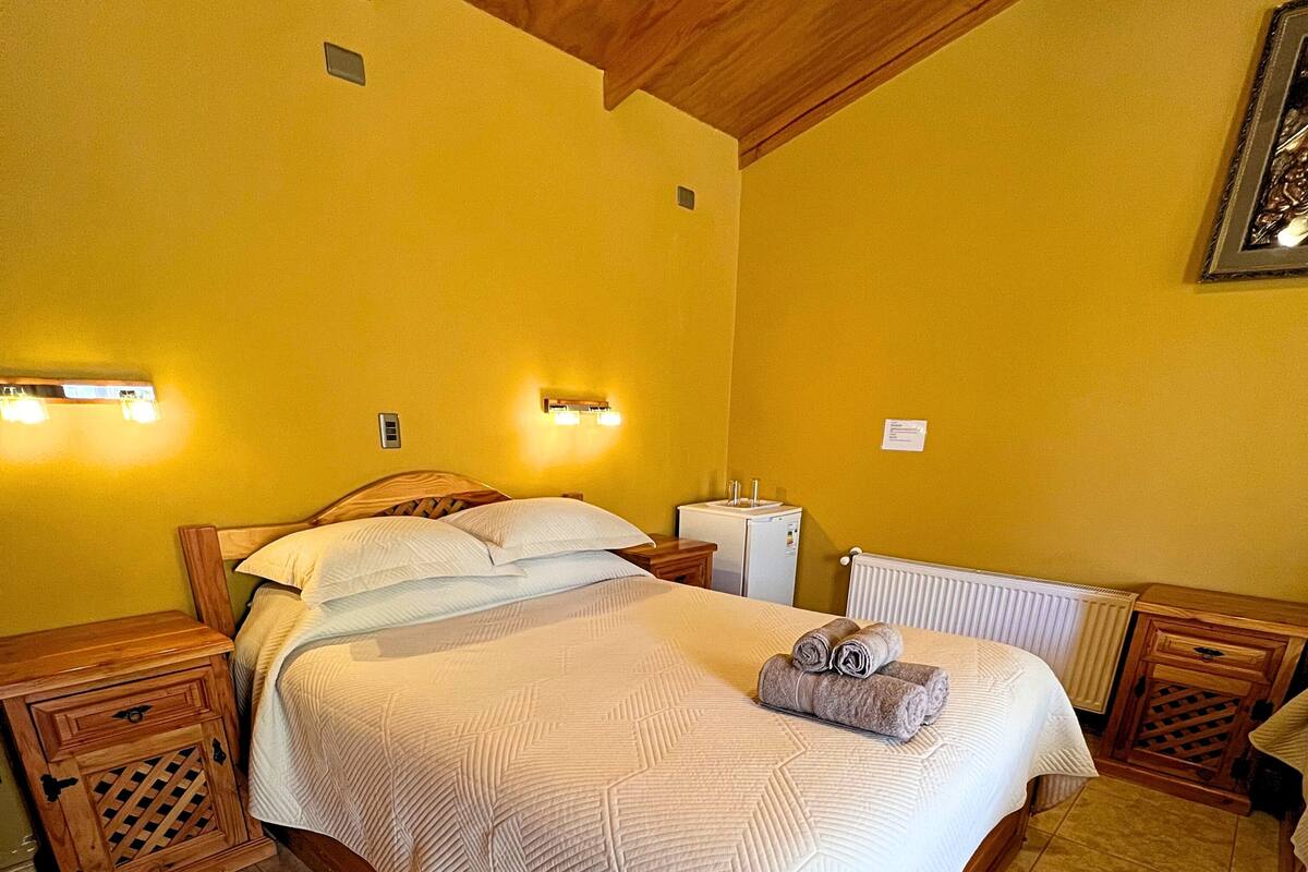Un lit bien fait, recouvert d’une couette blanche, une petite table de chevet, une lampe fixée au mur et un radiateur.