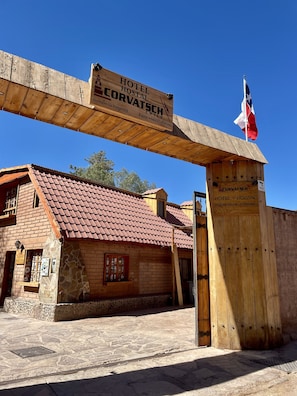 A wooden signboard for Hotel Hostal Corvatsch in Chile, a stone building with a tiled roof, and a flag.