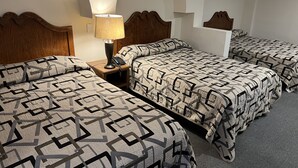 A hotel room with three beds, each with a geometric patterned bedspread. There is a wooden headboard on the left bed, a nightstand with a lamp and phone, and a small table with a lamp on the right bed. - Hotel Villas del Bosque (Aguascalientes)