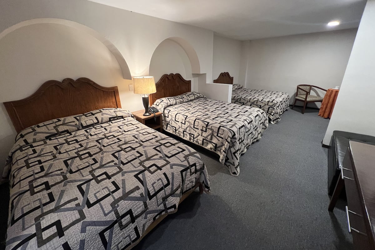 A hotel room with three beds, a wooden headboard, a desk with a lamp, and a chair.