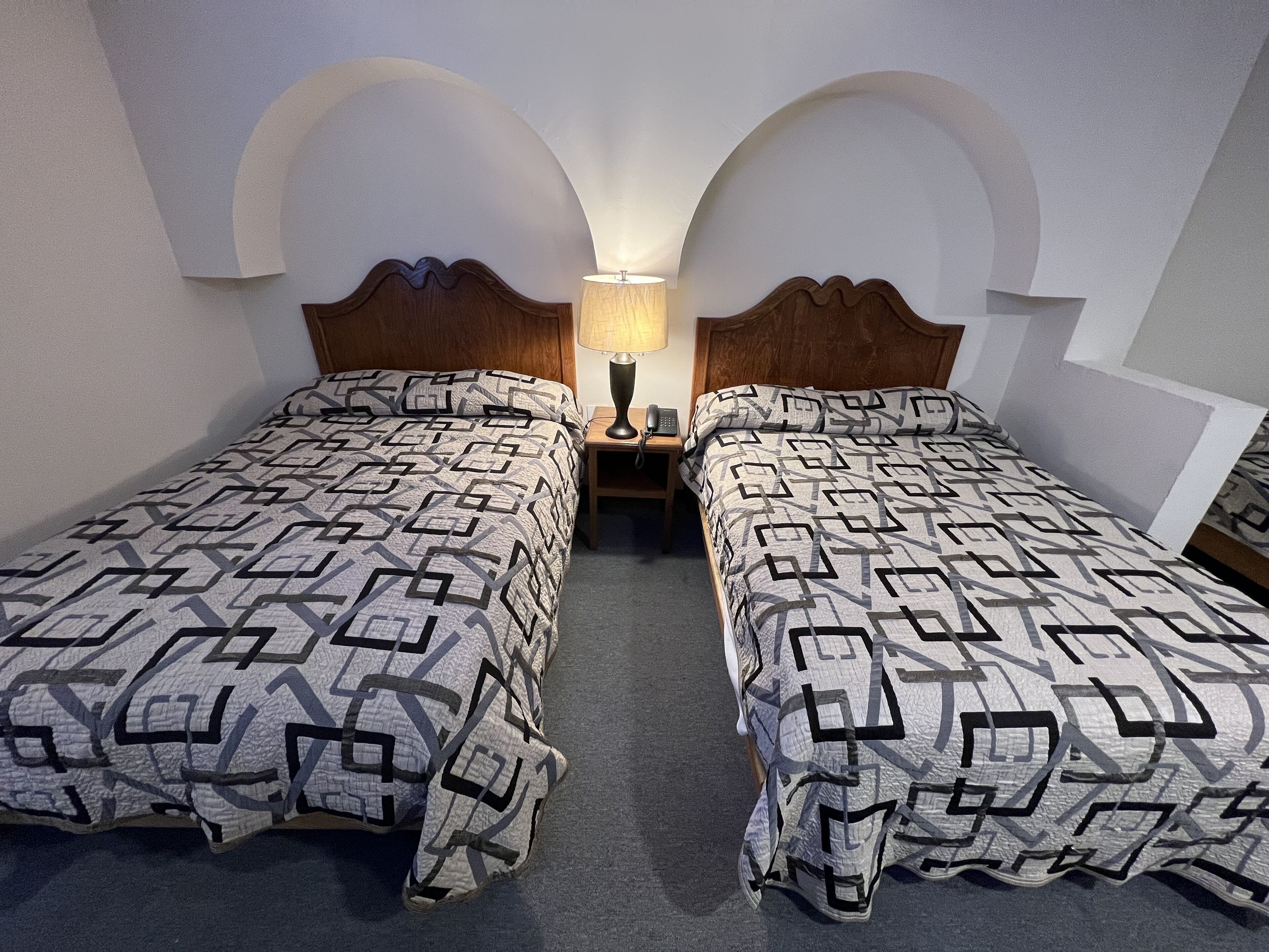A hotel room with two beds, each with a geometric patterned bedspread. There is a nightstand with a lamp and a telephone between the beds. The room has a simple, clean design with arched alcoves on the walls.