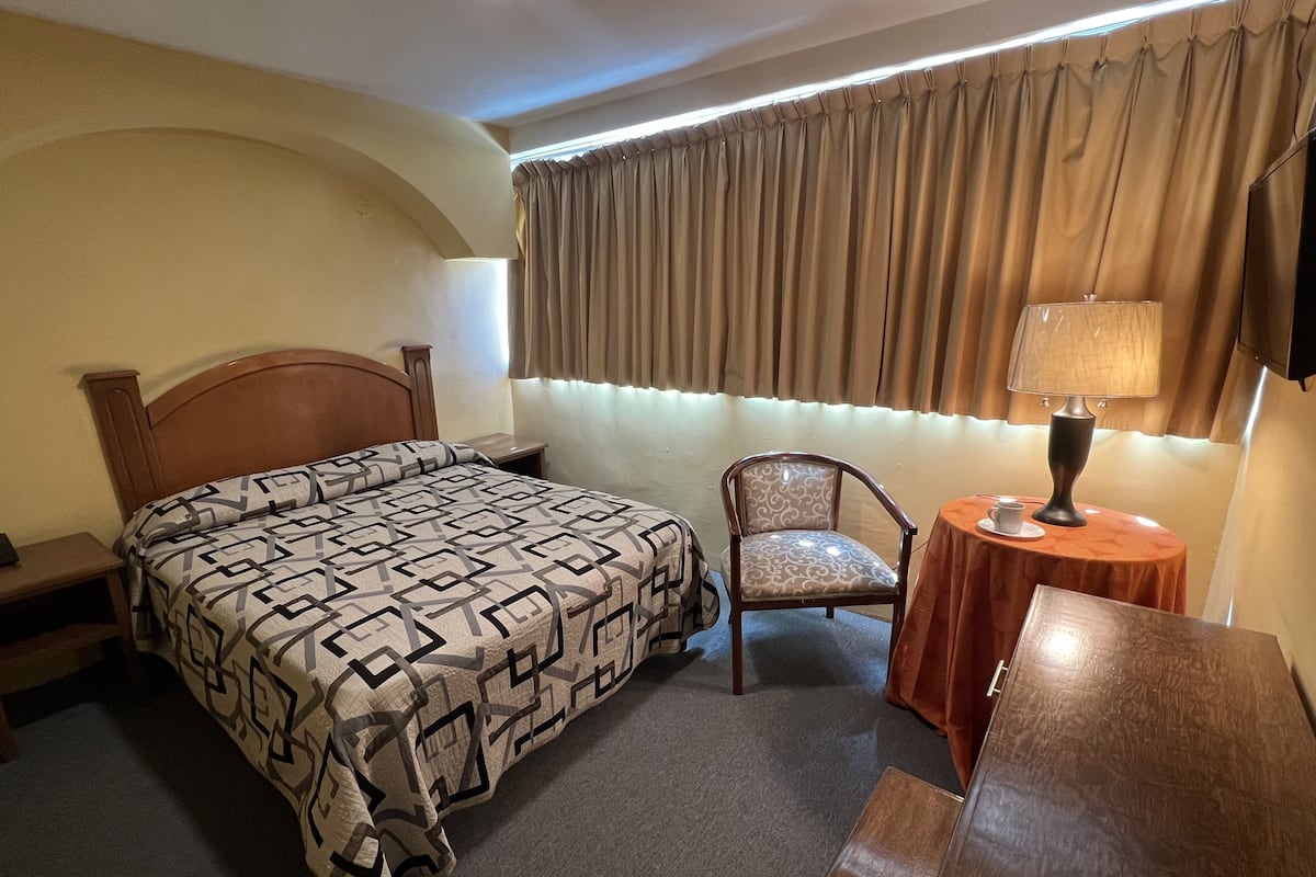 A hotel room with a bed, a chair, a small table with a lamp, and a TV.