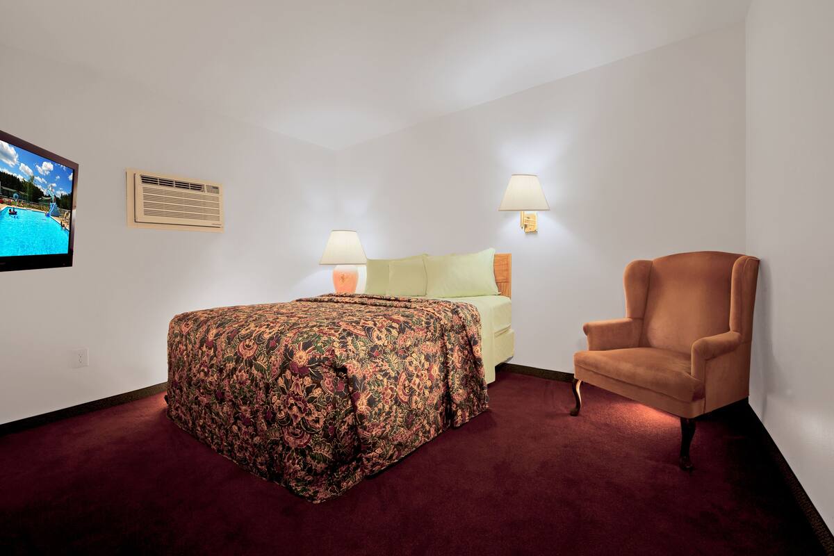 A hotel room with a bed, a chair, a TV, and wall-mounted lamps.